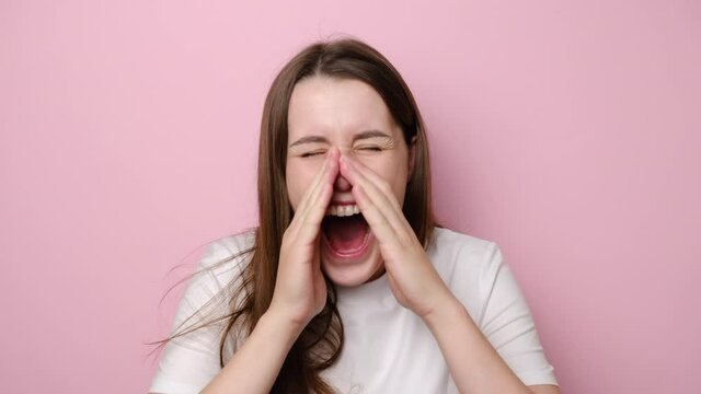 Portrait of desperate young annoyed angry woman shouting in megaphone hand gesture, expressing all hate aggression, screaming loudly, dressed in white t-shirt, isolated on pink studio background