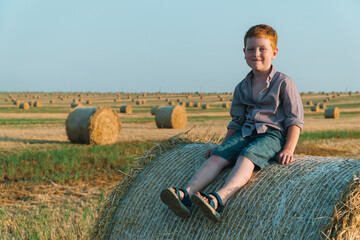 Naklejka na ściany i meble A red-haired boy sits on top of a straw bale on a wheat field