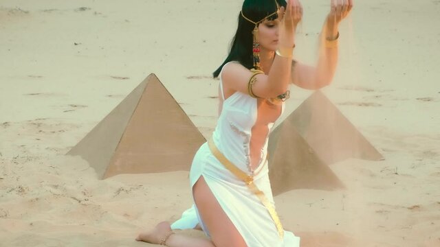 Beautiful sexy woman in image Egyptian pharaoh takes in hands yellow sand, plays. Queen Cleopatra Black hair wig bob cut Hairstyle. gold accessories jewelry necklace crown bracelets. Pyramids desert