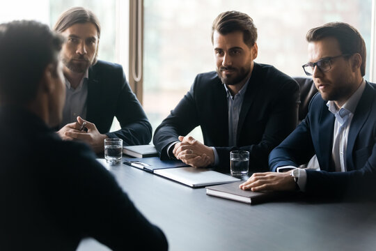 Focused young male group of employers in formal suits sitting at table with vacancy candidate, involved in job interview in office. Attentive confident hr managers listening to seeker at meeting.