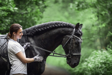 portrait of beautiful young woman standing near black friesian stallion in forest