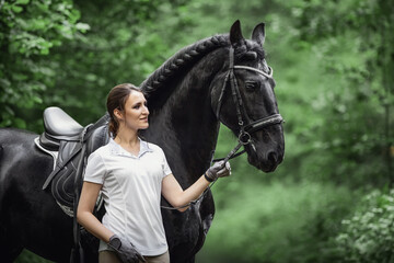 portrait of beautiful young woman standing near black friesian stallion in forest