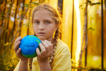 Fototapeta premium Portrait of freckled teenage girl holding planet in hands and looking at camera while standing outdoors in sunlight, copy space