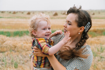 Beautiful young woman with fair-haired little son in her arms in the middle of a wheat field with bales in summer evening