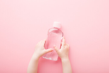 Baby girl little hands holding transparent plastic bottle of oil on light pink table background. Pastel color. Care about soft body skin. Close up. Point of view shot. Top down view.