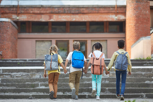 Full length back view at multi-ethnic group of children walking to school with backpacks and holding hands while walking up stairs to big building, copy space