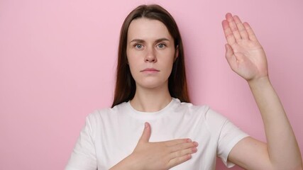 Portrait of sincere responsible young woman raising hand to promise, taking vow with serious dedicated expression, girl pledging allegiance, dressed in t-shirt,  isolated on pink studio background