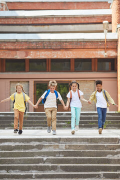 Vertical full length portrait of multi-ethnic group of children leaving school with backpacks and holding hands while running towards camera outdoors, copy space