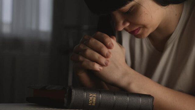 A woman prays with her hands crossed over a Bible for God's blessing in a dark room.