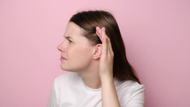 Confused young woman having hearing problems, asking to say louder, difficult to listen quiet talk, wears t-shirt, misunderstanding in communication, isolated on pink studio wall. Selective focus