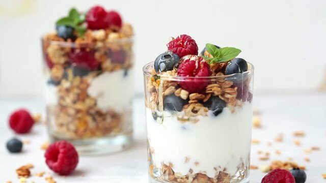 Pouring honey on yogurt parfait with granola, raspberries and blueberries in glasses, a light background. Healthy breakfast concept.