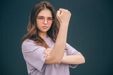 Young stylish trendy woman isolated over grey blue background. Serious strong powerful girl showing her strenght in arm by holding one of them up. Modern stylish girl look straight.