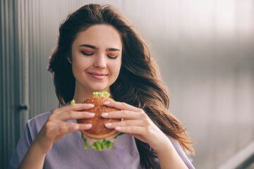Young stylish trendy woman isolated over grey blue background. Happy positive girl looking at tasty delicious burger in hands. Unhealthy but yummy meal.