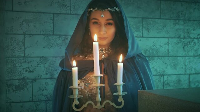 Beautiful mystic woman ghost walks at night in dark castle, holding candlestick with burning candles. spirit of medieval queen in blue dress, hood cape, gothic clothing. Horror art concept halloween