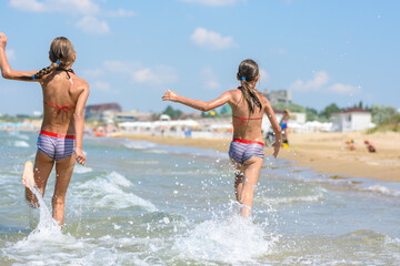 Two girls joyfully run along the sea on a warm summer day, rear view
