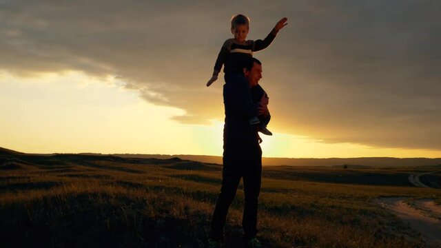 Silhouette of a happy family at sunset. The son sits on the shoulders of his father and depicts the flight of an airplane. The family is relaxing in the fresh air, dreaming of traveling.