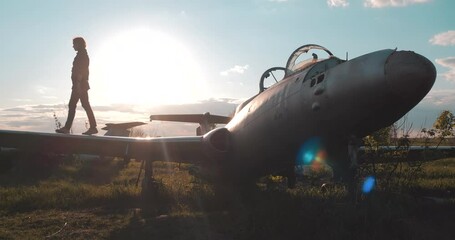 Medium view of tourist girl walking along wing of broken Soviet combat fighter plane, then turning around and throwing head back from enjoying on airfield near historical transport, background of sun.