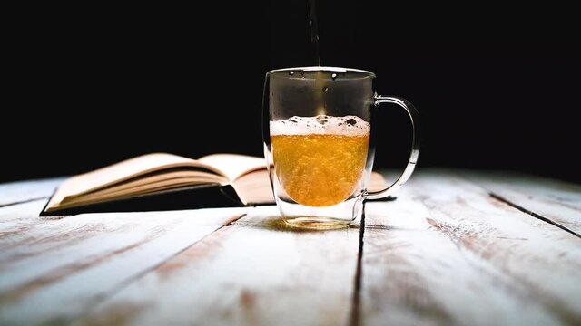 Close up of a glass cup being filled with golden tea with an open book behind on a wooden table. Relaxation and lifestyle concept.