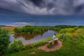 Summer landscape with river and person looking towards the storm clouds