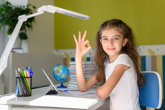 Girl sitting at a desk with textbooks and a tablet. girl looking at camera, smiling and showing ok sign