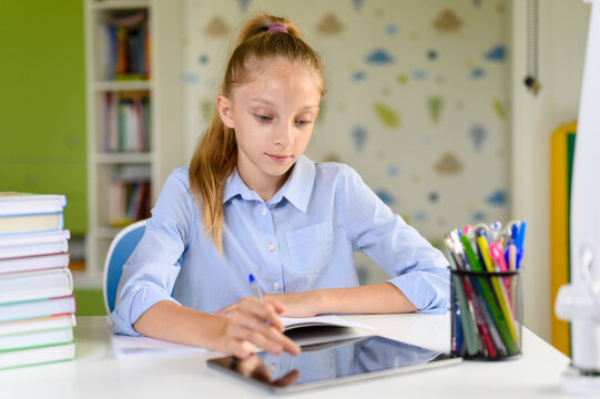The girl does homework and uses the tablet. Girl schoolgirl with long blonde hair sits at a desk at home.