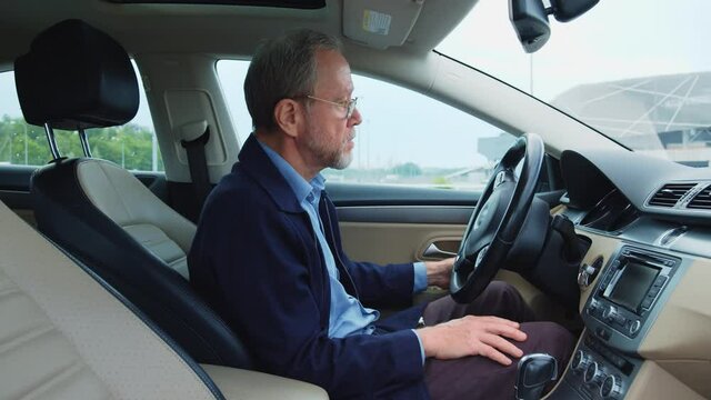 Caucasian bearded wealthy man sits inside his modern automobile on parking space. Profile handsome grey-haired old man starting a car going to work. Business people. Transportation.