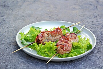 Grilled pork with bacon on wooden skewers, on a white ceramic tray on a gray concrete background. Served with lettuce.