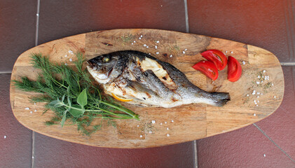 Dorado fish was cooked with lemon over an open fire and lies on a wooden Board with herbs and slices of tomatoes. The concept of cooking healthy and delicious food.