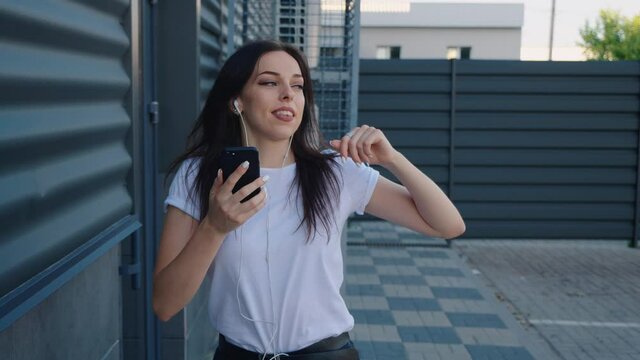 Portrait of smiling young woman dancer listens music with smartphone on a urban background. Pretty girl student holds smart phone in hands and enjoys the music with earphones and dancing in a city