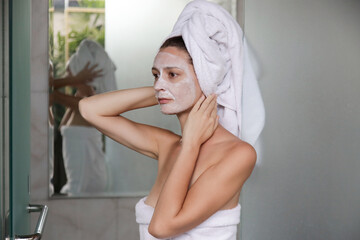 Young woman in bathroom wrapped in towels with facial mask. Home spa and skin care concept.