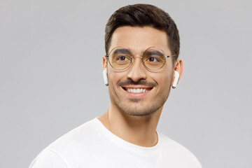 Close-up portrait of young handsome man in white t-shirt, wearing eyeglasses and wireless earphones, making plans and dreaming with smile, isolated on gray background