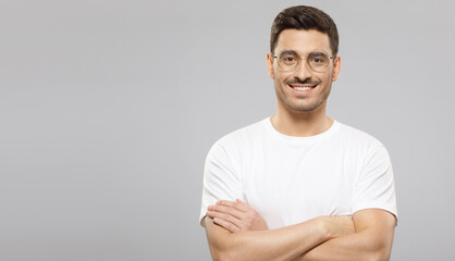 Horizontal banner of young smiling handsome man in white t-shirt and round glasses, standing with crossed arms, isolated on gray background with copy space