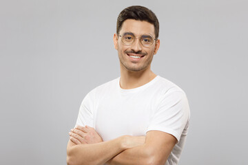Portrait of handsome smiling guy dressed in white t-shirt, wearing round eyeglasses, holding arms crossed, ready to help and consult, isolated on gray background