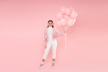 Full length portrait of happy girl standing in winter outfit and colored glasses, holding bunch of birthday balloons, isolated on pink background