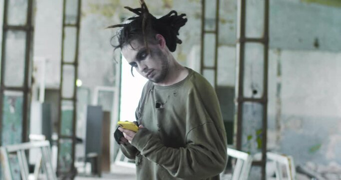 Frightening dire zombie man with dreadlocks walking around the abandoned building. Pale infected living dead scrolling the news feed on the smartphone. Halloween, costume party, supernatural concept.