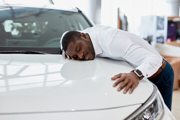 african american man hugs new car he dreams about, he stand in cars showroom next to white...
