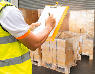 Warehouse worker writing on paper clipboard, his checking order shipment boxes on pallets. Warehouse inventory management.