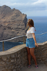 A long-legged girl stands half-turned on the observation deck of Mirador Es Colomer and looks at the sea, her hair covering her face. Cape de Formentor. Majorca. Spain.
