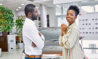 pretty african american female asks her husband to buy a car, she looks at camera, man look at her and think. isolated in car showroom