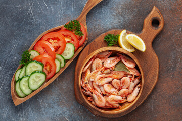 Boiled shrimp prawns in a shell with lemon and vegetables in a plate on a wooden background. Top view