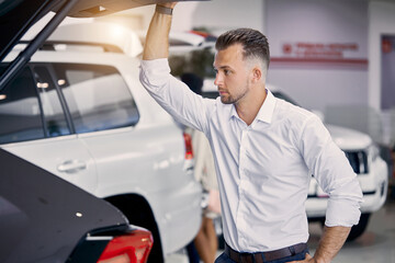 handsome caucasian guy examine the trunk of car in dealership , he looks inside of car from back and think buy it or not