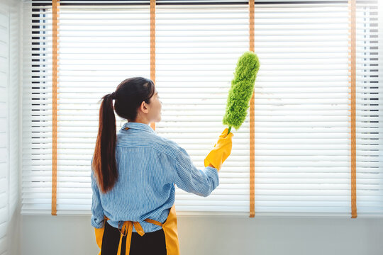 Young Asian Woman Cleaning House Wiping Dust Using Feather Broom And Duster While Cleaning On Window House Keeping Concept