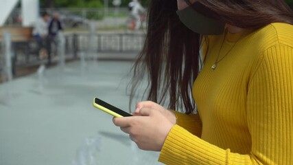 Front view of a mixed race woman with long dark hair out and about in the city streets during the day, wearing a face mask against air pollution and coronavirus Covid19, using a smartphone - Powered by Adobe