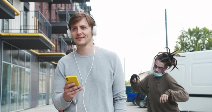 Dire male zombies chasing man to eat him on the street. Young guy in headphones walking, listening to music unaware of hungry living dead following him. Apocolypse, halloween, supernatural concept.
