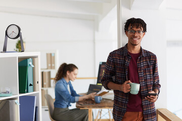 Fototapeta premium Waist up portrait of smiling African-American man holding mug and looking at camera while standing against wall in contemporary office interior, copy space