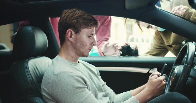 Young Caucasian man sitting in the car scrolling news feed on a smartphone, tapping message. Frightening zombies tries to get inside the car to kill the guy. Apocalypse, supernatural, evil concept.