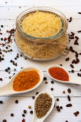 Ingredients for cooking pilaf: rice in a glass jar in the background, garlic, cumin, saffron and paprika in white spoons, barberry and black pepper are scattered on the table in the foreground.