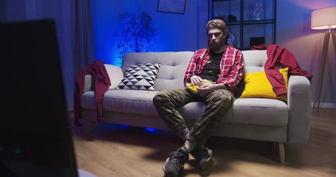 Scary evil male zombie sitting on sofa in the house. Pale infected undead man eating chips, crisps, snacks at the Halloween party. Supernatural, walking dead, afterlife, apocalypse concept.