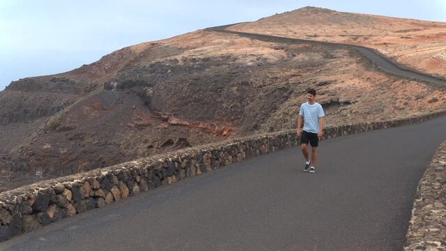 Hombre joven camina por una carretera y sube a un muro para observar la isla de La Graciosa. Lanzarote. Islas Canarias
