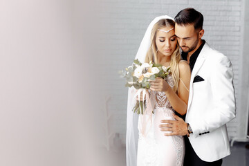 wedding couple groom and bride posing in a white studio. White dress. Blonde bride with bouquet in her hands. White backgroud.